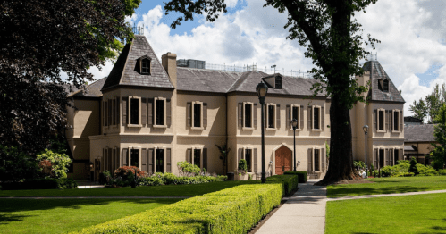 A large, elegant building with a landscaped garden, featuring tall windows and a symmetrical design under a blue sky.