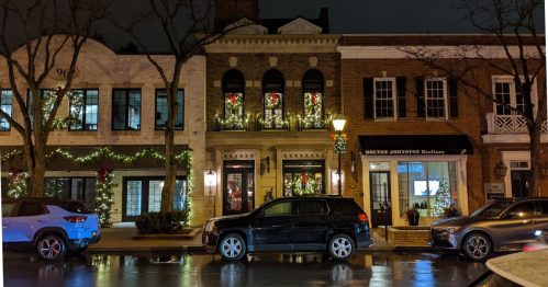 A street view of festive buildings adorned with holiday lights and decorations at night, with parked cars in front.
