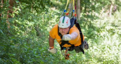 A person in an orange shirt and helmet zip-lining through a lush green forest.