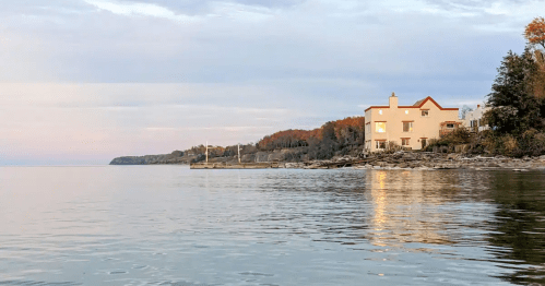 A calm lake scene at sunset, featuring a pink house and trees with autumn foliage along the shoreline.