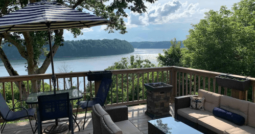 A scenic deck with outdoor furniture overlooks a calm lake surrounded by trees and a blue sky with clouds.