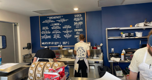 Two chefs working in a kitchen with a menu board on the wall and food preparation areas visible.