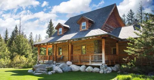 A rustic wooden cabin with a porch, surrounded by trees and large rocks under a blue sky with fluffy clouds.