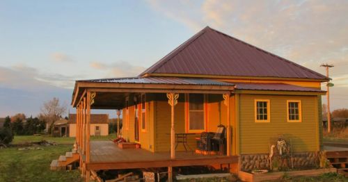 A colorful yellow house with a red metal roof, featuring a spacious porch and surrounded by a grassy landscape at sunset.