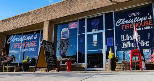 Exterior of Christine's Firehouse Bar & Grill, featuring large windows, a sign for live music, and outdoor seating.