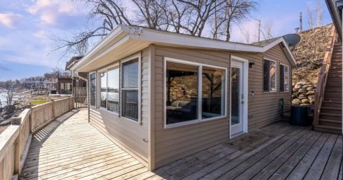 A cozy lakeside cabin with large windows, wooden deck, and a staircase leading up the hill.