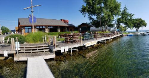 A waterfront restaurant with outdoor seating, surrounded by trees and a clear blue sky, next to a calm lake.