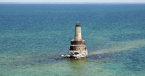 A tall lighthouse stands on a rocky base in calm blue waters, surrounded by a gradient of turquoise and deep blue.