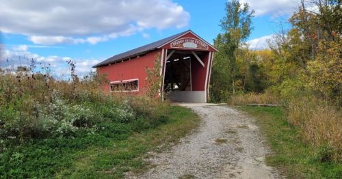 A red covered bridge stands at the end of a gravel path, surrounded by greenery and trees under a cloudy sky.