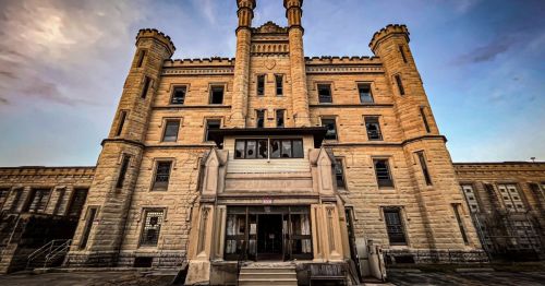 Historic stone building with towers, featuring large front entrance and windows, set against a cloudy sky.