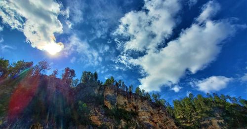 A vibrant sky with fluffy clouds above a rocky cliff and lush green trees, illuminated by sunlight.