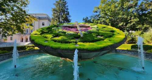 A large floral clock surrounded by water fountains and greenery, set against a clear blue sky.