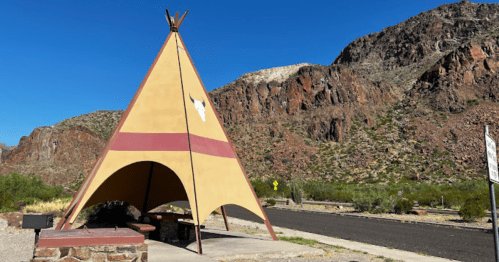 A colorful teepee structure near a road, set against rocky mountains and clear blue skies.