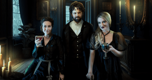 Three elegantly dressed individuals stand in a dimly lit room, each holding a drink, with candles and dark decor around them.