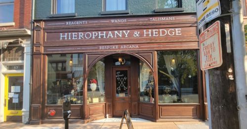 A storefront with the sign "Hierophany & Hedge" featuring wooden accents and a mystical theme.