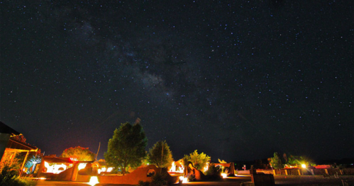 A starry night sky over a desert landscape, with illuminated structures and trees in the foreground.