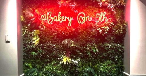 A vibrant green wall with various plants and a neon sign that reads "Bakery On 5th."