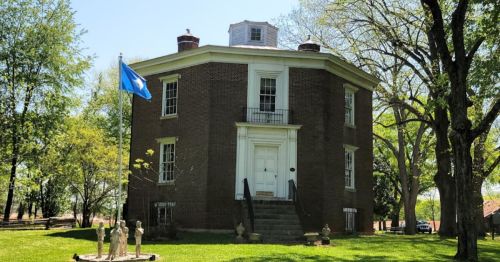 A historic round brick building with a flag, surrounded by trees and sculptures in a grassy area.