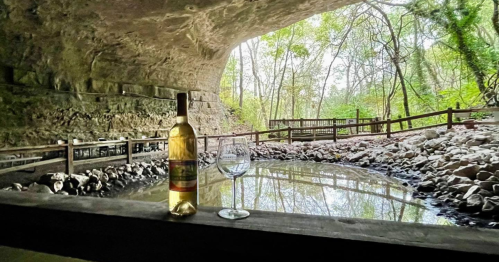 A bottle of white wine and a glass sit on a wooden table by a serene cave entrance with a pond and trees in the background.