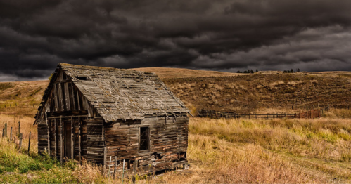 A weathered, abandoned wooden cabin in a grassy field under a dark, stormy sky.