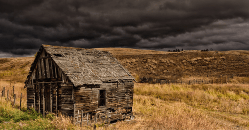 A weathered, abandoned wooden cabin in a grassy field under a dark, stormy sky.