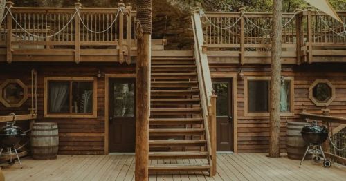 A wooden cabin with a staircase, surrounded by trees, featuring outdoor grills and a rustic design.