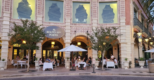 Outdoor dining scene at a restaurant with elegant architecture, featuring tables under umbrellas and guests enjoying their meals.