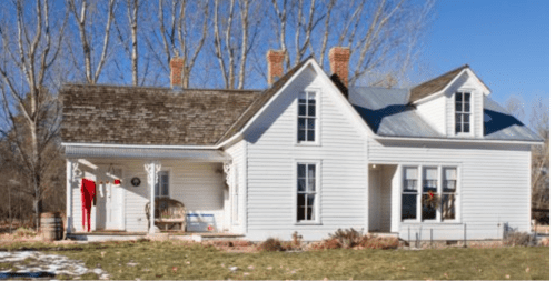 A charming white house with a sloped roof, surrounded by trees and a grassy yard, featuring a porch and holiday decorations.
