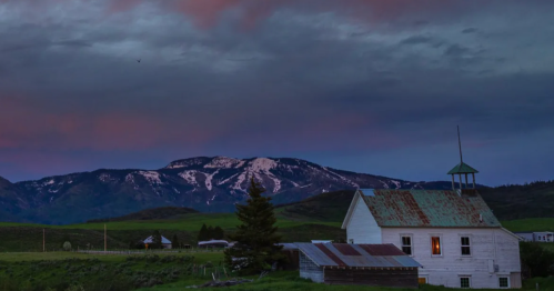A rustic white house with a green roof sits in a valley, surrounded by mountains and a colorful sunset sky.