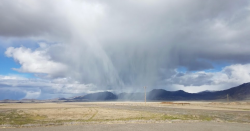 A dramatic sky with dark clouds and rain falling over a mountainous landscape, with open fields in the foreground.