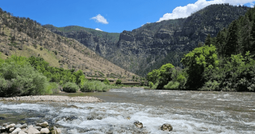 A scenic river flows through a lush valley surrounded by mountains and greenery under a clear blue sky.
