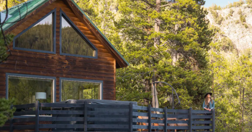 A wooden cabin surrounded by trees, featuring large windows and a deck with a person sitting nearby.