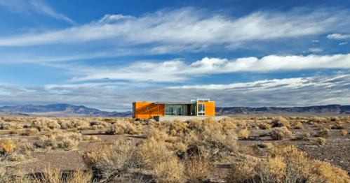 A modern orange house stands alone in a vast desert landscape under a blue sky with scattered clouds.