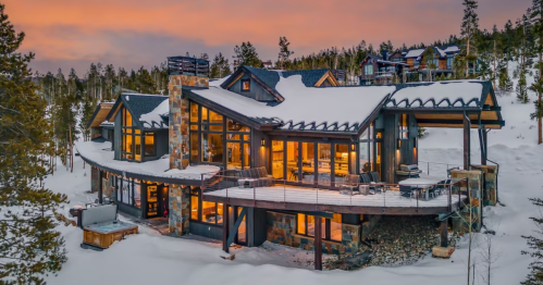 A modern mountain home with large windows, surrounded by snow and trees, illuminated at sunset.