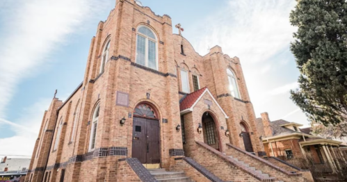 A large brick church with arched windows and a red roof, surrounded by trees and a clear blue sky.