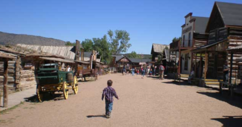A child walks down a dusty street lined with wooden buildings and wagons in a historic western town.