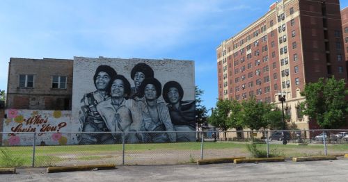 A mural of five men smiling, set against a brick wall, with a nearby building and greenery in the foreground.