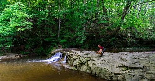 A person sits on a rocky riverbank beside a flowing stream, surrounded by lush green trees.
