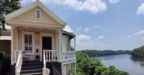 A charming house with a porch overlooking a calm river under a blue sky with fluffy clouds.