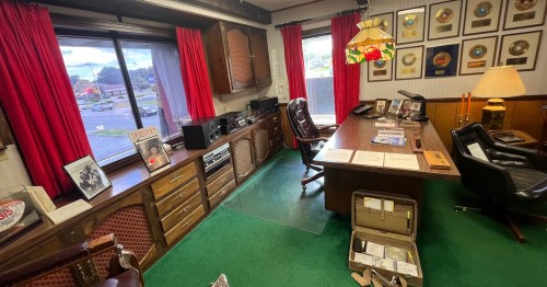 A vintage office with a wooden desk, red curtains, awards on the wall, and a green carpet, featuring a record player and memorabilia.