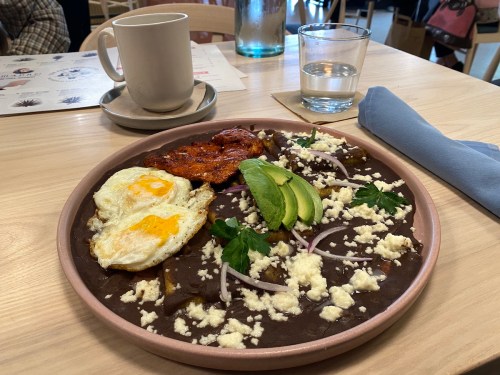 A plate of huevos rancheros with eggs, avocado, cheese, and a side of chicken, served with coffee and water.
