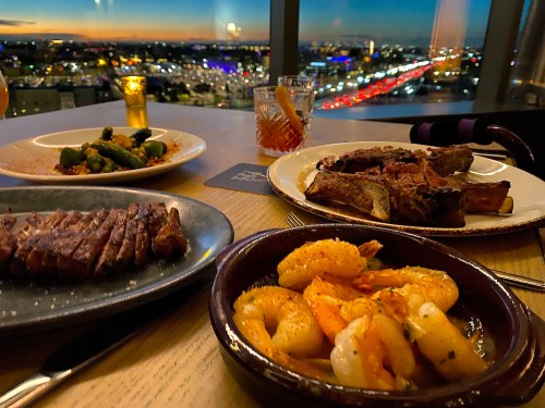A table with gourmet dishes: grilled shrimp, steak, and ribs, with a city skyline view at sunset in the background.