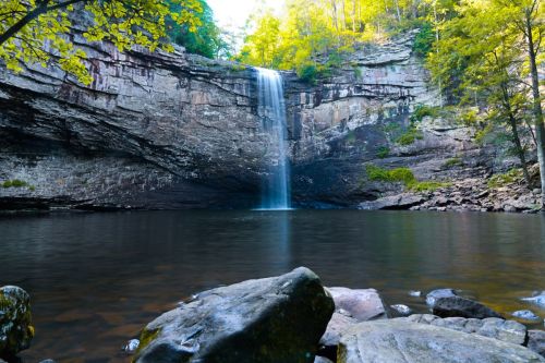 A serene waterfall cascades into a calm pool, surrounded by lush greenery and rocky terrain.