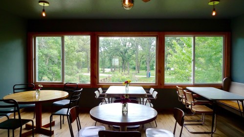 Cozy dining area with wooden tables, chairs, and large windows overlooking a green outdoor space.