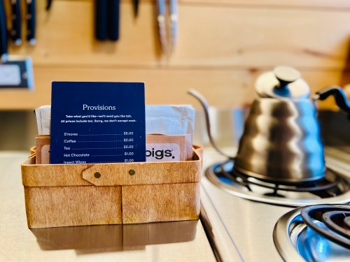 A wooden basket holds a menu and various drink options, with a kettle on a stove in the background.