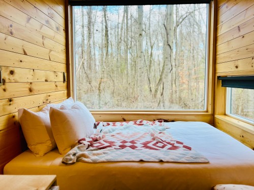 Cozy bedroom with a large window overlooking a forest, featuring a bed with white pillows and a patterned blanket.