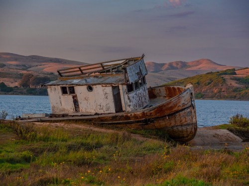 An abandoned, weathered boat rests on a grassy shore with hills and a calm body of water in the background at sunset.