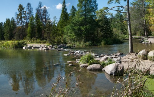A serene river scene with smooth rocks, lush greenery, and trees reflecting in the calm water under a clear blue sky.