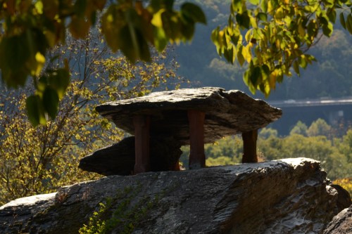 A large, flat rock balanced on smaller stones, surrounded by trees and a scenic landscape in the background.