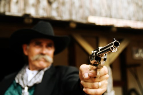 A man in a cowboy hat points a revolver towards the camera, with a blurred background of a rustic building.